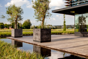 Duurzaam houten terras omgeven door waterpartij en siergras zodat er een mooi terras ontstaat in strakke lijnen welke dit moderne nieuwbouw bedrijfspand ondersteunen in het geheel te Oudewater